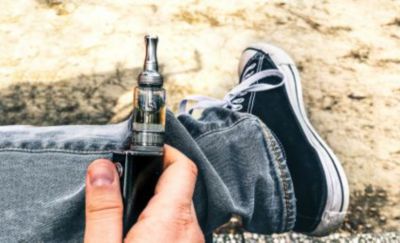 A close-up of a person sitting crossed legged, holding thier vape in their hand while resting it on their left leg