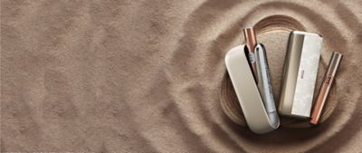 Dve napravi iz serije IQOS ILUMA i MINERA – ILUMA i ter ILUMA PRIME i – ter držala in žepna polnilnika na peščeni podlagi.