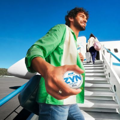 Muž v zelenej košeli nastupujúci do lietadla a v ruke drží škatuľku ZYN.