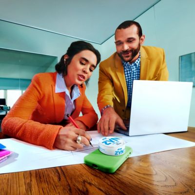 Žena píšuca na papier nad ňou stojí muž a pred sebou má položený telefón, na ktorom leží škatuľka ZYN.