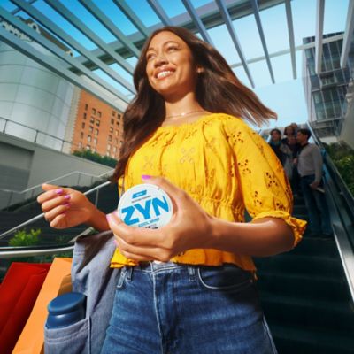 Žena na eskalátore s nákupnými taškami a škatuľkou ZYN v ruke.