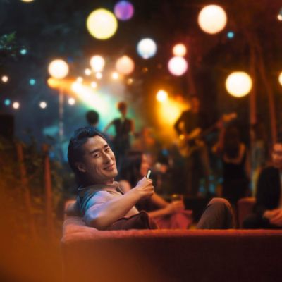 A person is sitting on a couch in a dimly lit room with colorful lights and hanging lanterns. There are several people in the background. The atmosphere suggests a relaxed, social setting.