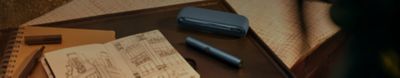 A cozy setting with a notebook open to a page containing architectural sketches, placed on a black tray. There are also two brown pencils, a closed brown notebook, and a blue ILUMA holder and charger next to it.
