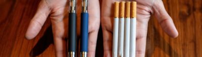 An adult holding two vape devices in one hand and four traditional cigarettes in the other, comparing vaping and smoking.