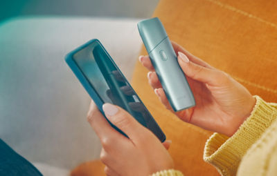 Hands holding a mobile phone and IQOS ORIGINALS ONE device.